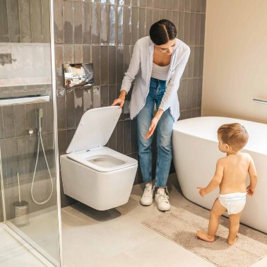 A parent gestures to the toilet as a child in a nappy backs away, uncertain